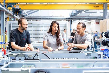 3 workers standing in factory hall discussing in front of an industrial device.