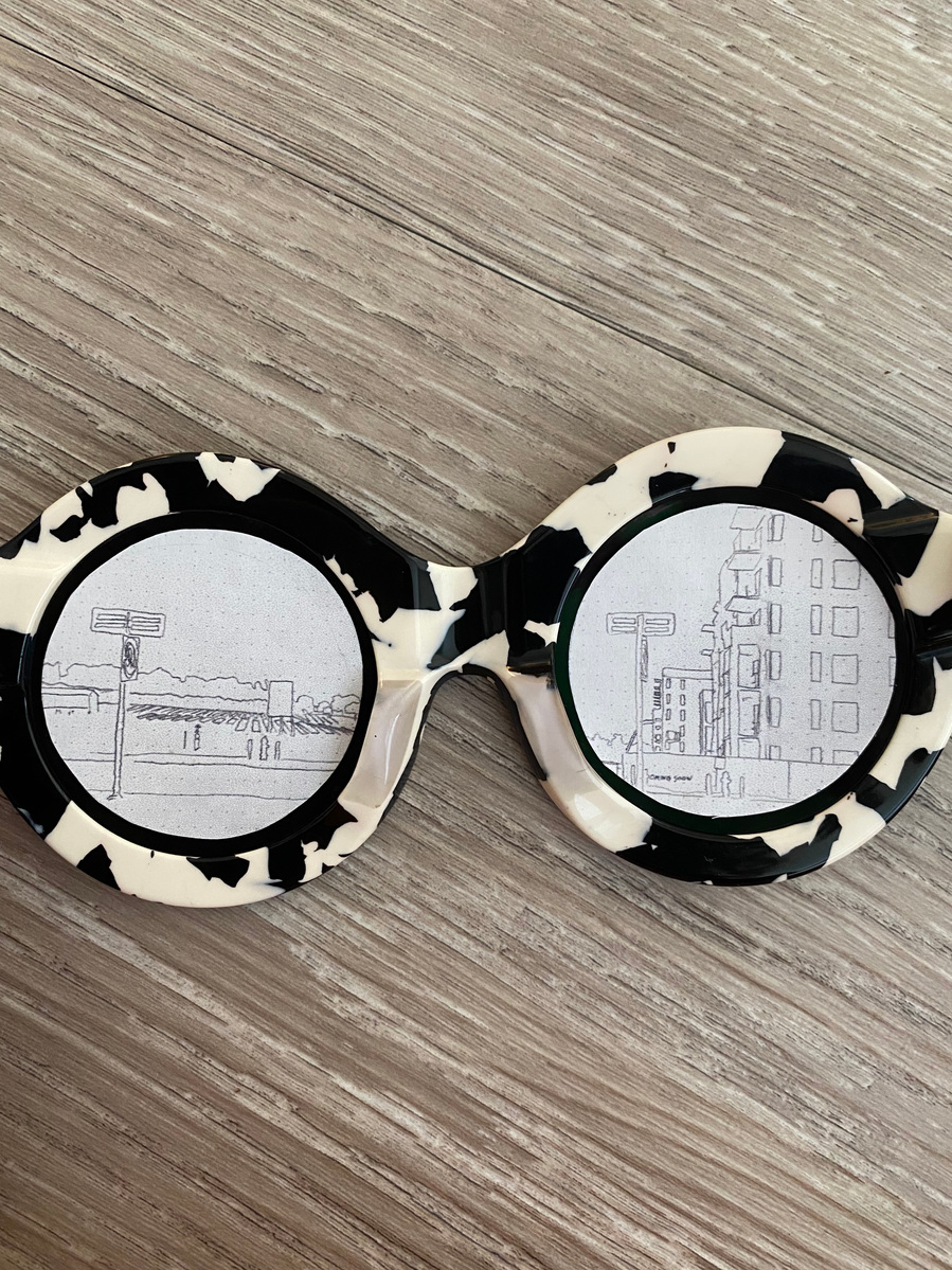 inside of sunglasses with paper circles on the eyes. Left eye has a drawing of strawberry fields. Right eye has a drawing of a building in the same field