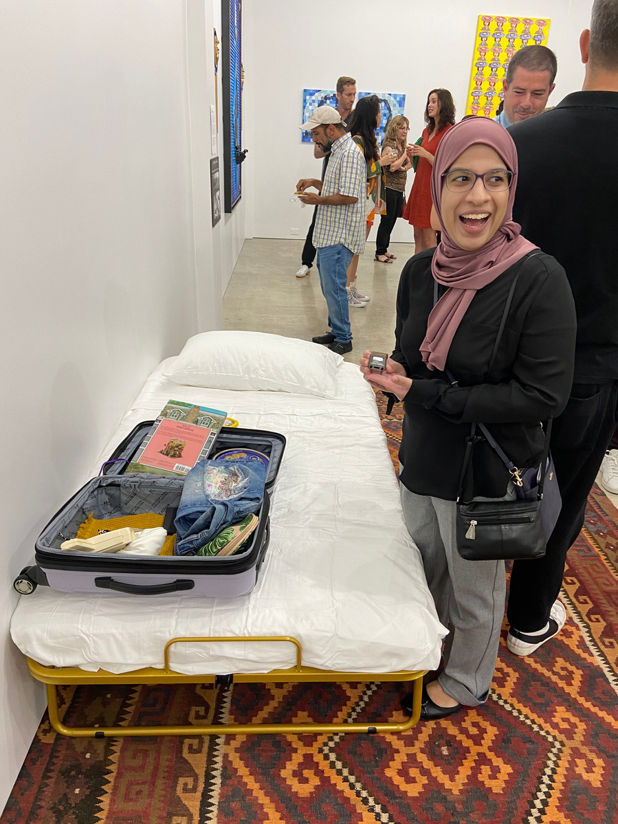 a woman with a hijab stands in front of the bed and holds a working tiny tv in between her fingers. She looks to someone off camera to the right with a face or surprise and delight