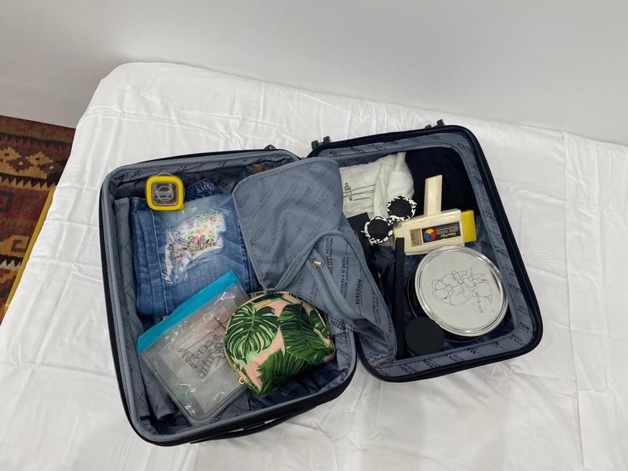 photo looking down into an open suitcase on a bed. Items in suitcase include Fisher Price movie viewer, jeans with a large hole on the knee, a ziplock bag, sunglasses, a toiletry bag