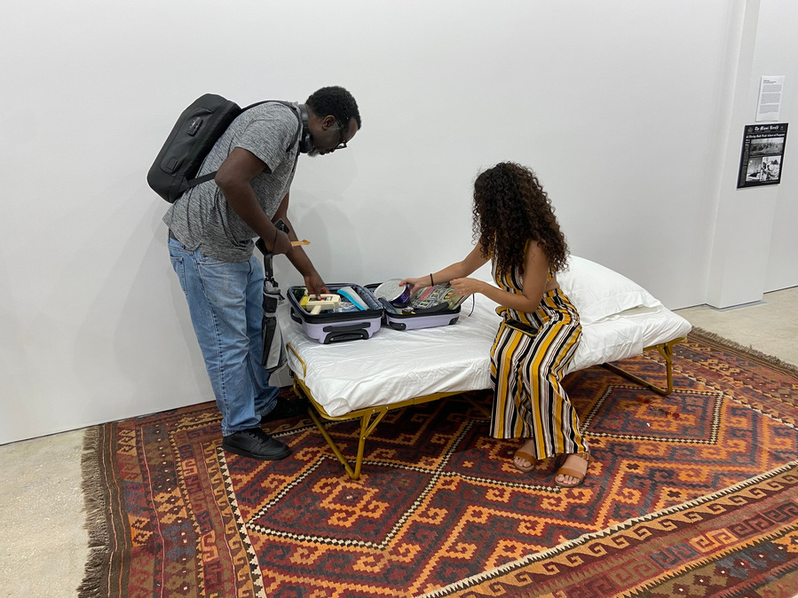 Photo of a black man with a backpack leaning over and looking at items in the suitcase while a curly haired woman sits on the bed grabbing for something in the suitcase