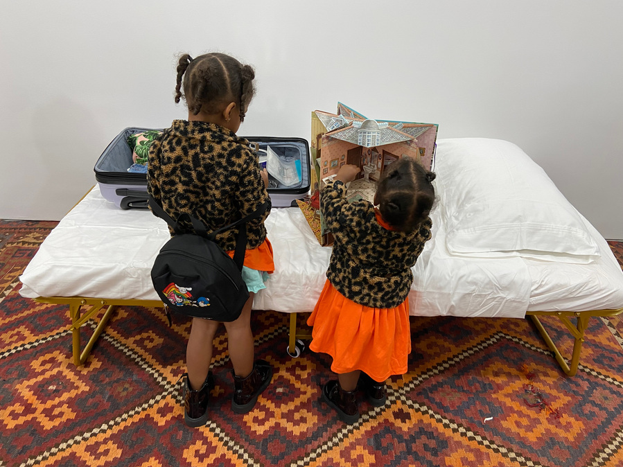 photo of two little black girls standing in front of the bed. The younger one on the right holds a pop up dollhouse book open in her hands while her sister looks at the dollhouse