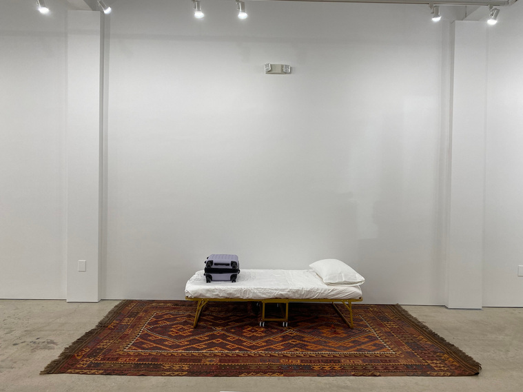 Photo of a bed against a white wall (lengthwise) sitting on a large brown and orange rug with geometric designs. The bed has white sheets, one white pillow on the right end and a closed purple suitcase on the left. 