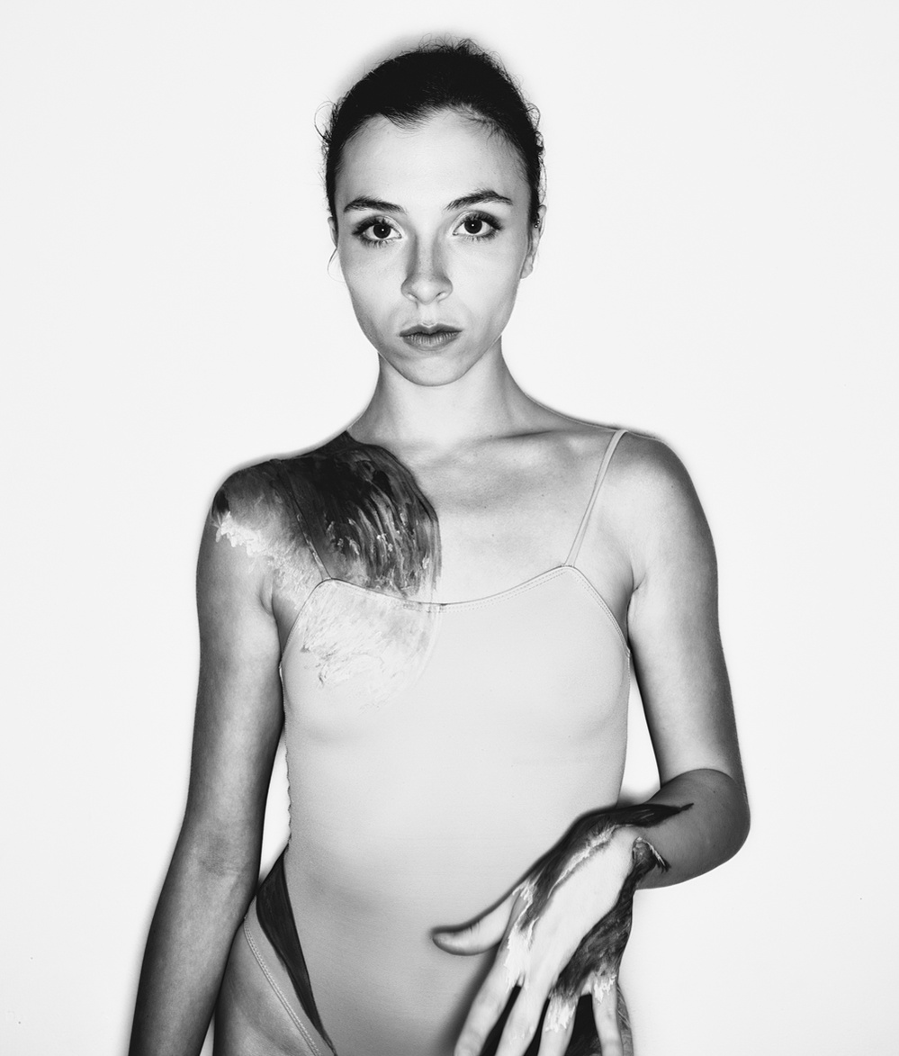 Black and white photograph of a female ballet dancer looking into the camera with body paint of waves on her shoulder and forearm.