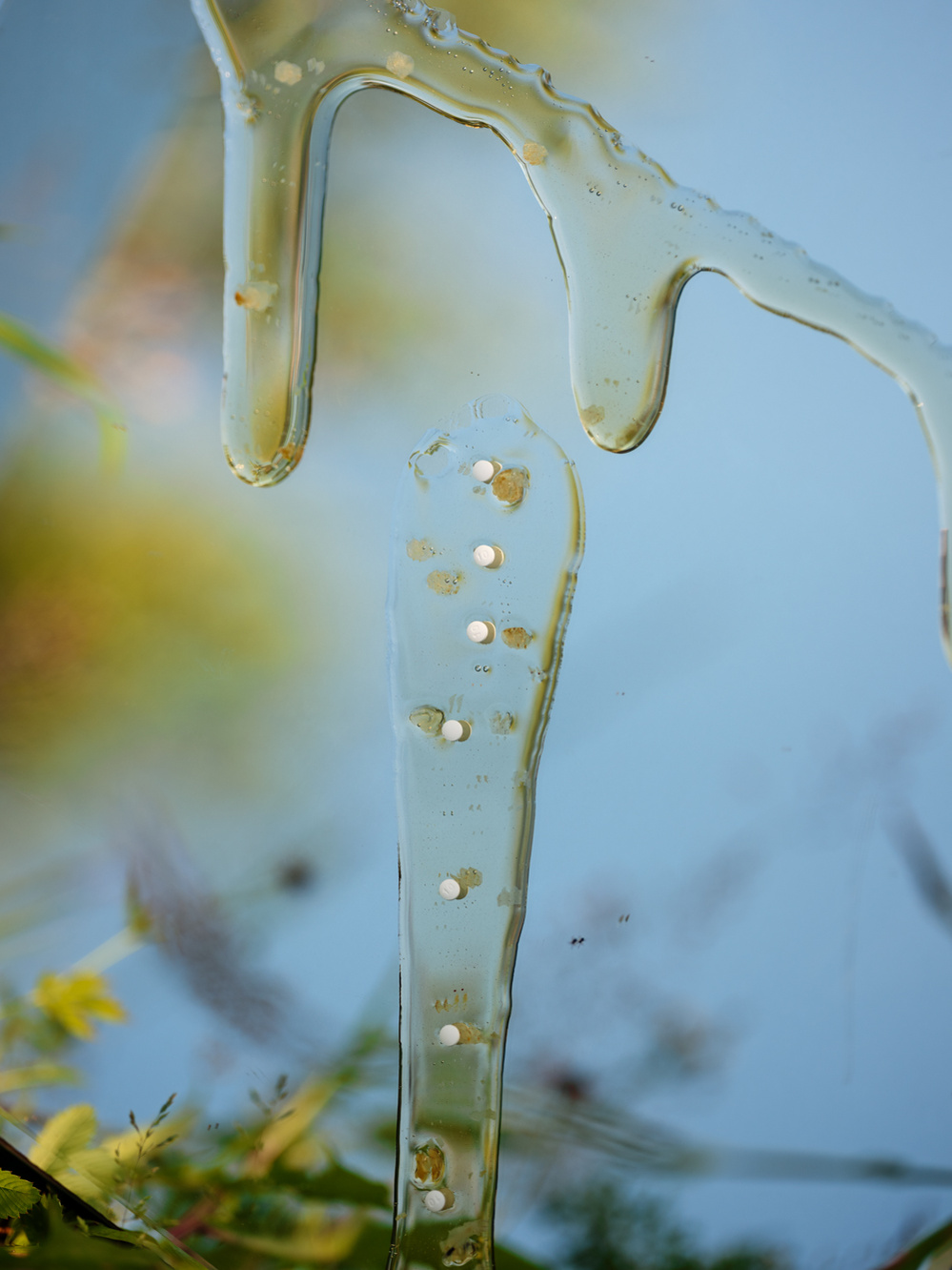 Photograph of pills in honey trickling down a mirrored surface with blue sky reflected in the mirror.