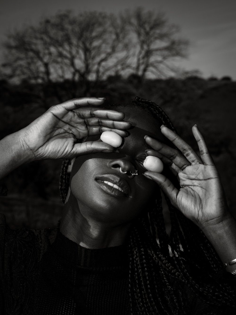 Black and white photograph of a model holding small fruits over her eyes with trees in the background.