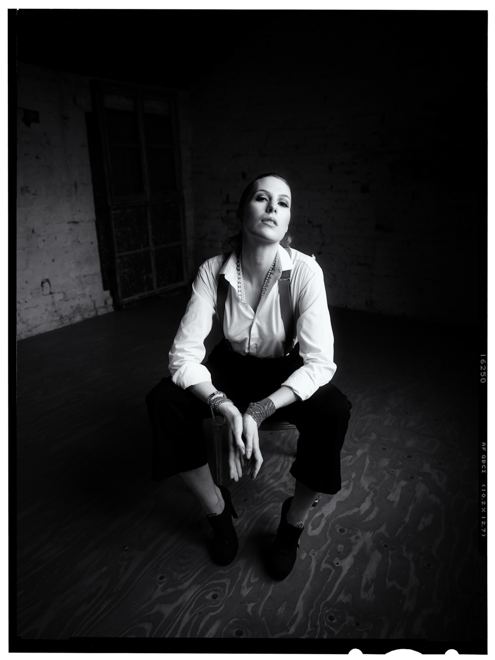 Black and white photograph of a female model sitting in a darkened room wearing a white shirt and braces and looking into the camera.