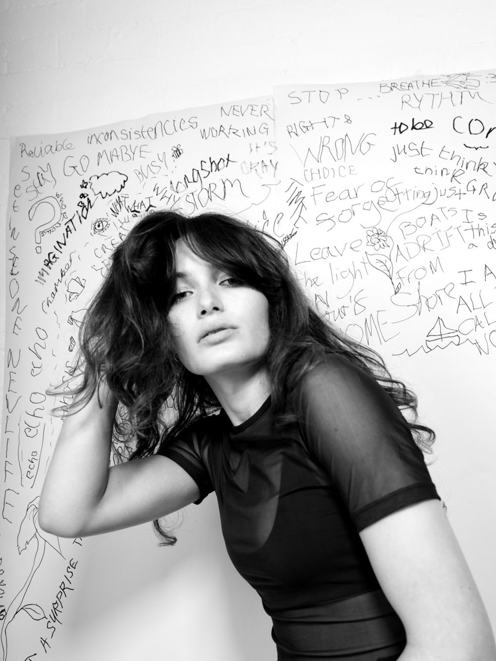 Black and white portrait of a female model with a hand in her hair looking at the camera with a wall covered with writing behind her.