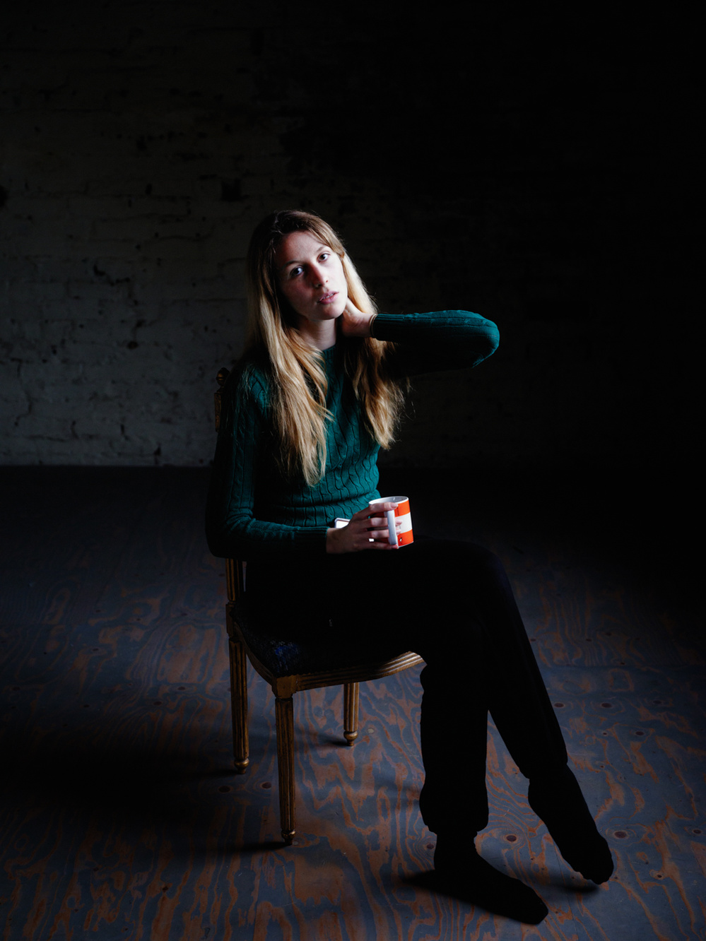 Colour portrait photograph of a female model in a green jumper sitting in a darkened room looking at the camera and holding a mug of tea in one hand.