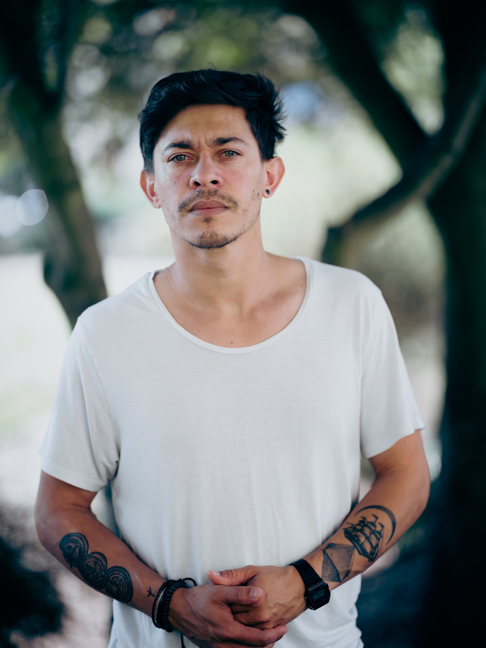 Colour portrait photograph of a male model wearing a white t-shirt looking at the camera with trees in the background.