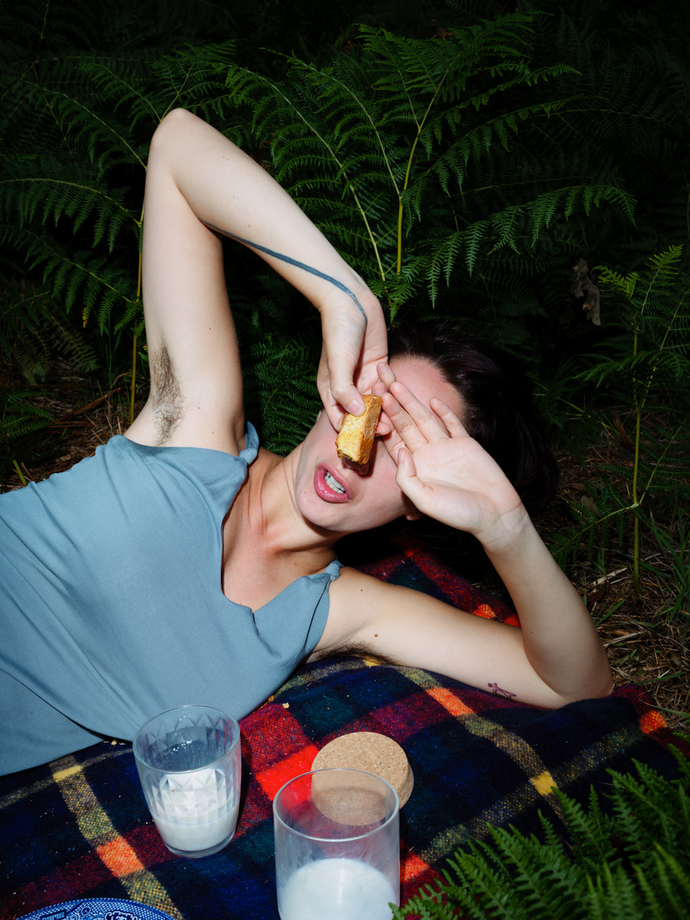 Colour photograph of a woman in a grey dress lying on a picnic rug with her hands over her eyes and holding a biscuit in one hand.