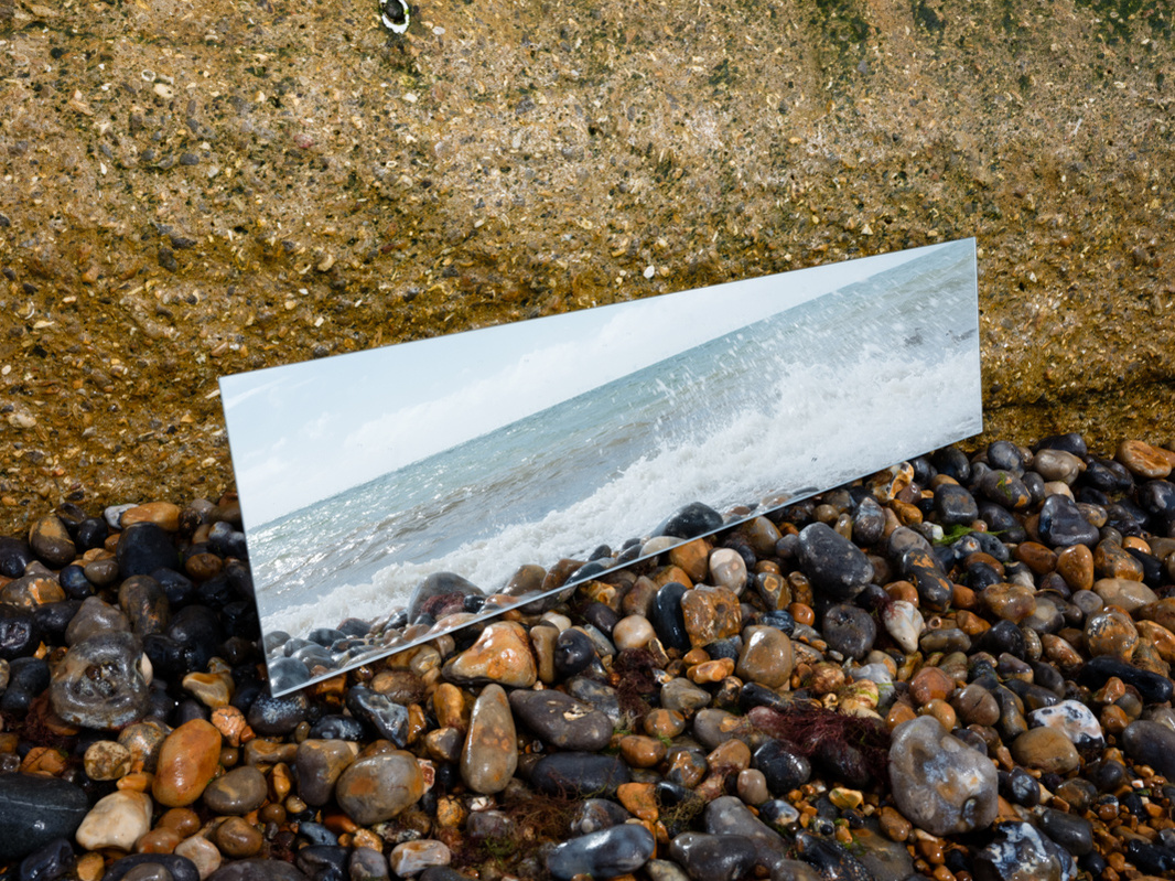 Colour photograph of a long mirror set against rocks on a pebble beach reflecting sea and waves.