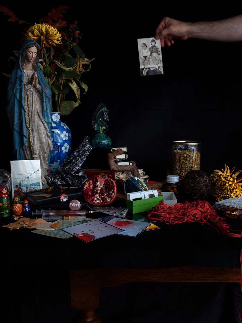 Colour photograph of a collection of personal items on a tabletop with an arm in the frame holding a black and white photograph of two boys.