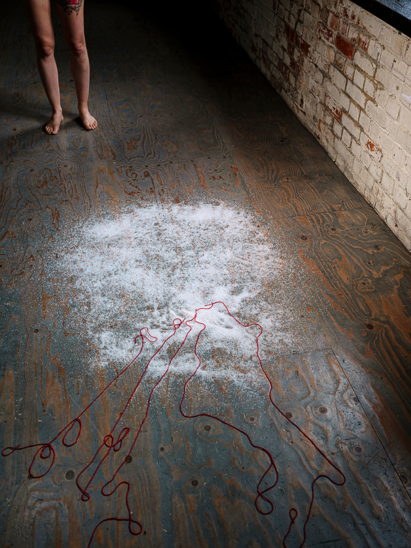 Colour photograph of salt spilt on a wooden floor with some red thread laid out in the salt and a pair of legs in the background.