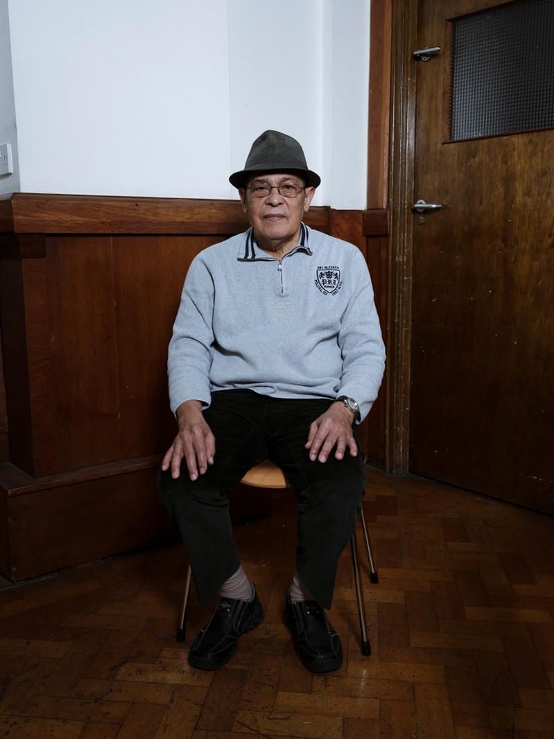 Colour photograph of an elderly black man with a trilby on his head sitting in a room holding looking straight into the camera.