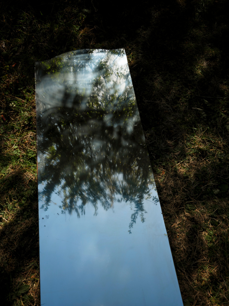Colour photograph of a mirror lying on the ground reflecting trees and a blue sky.
