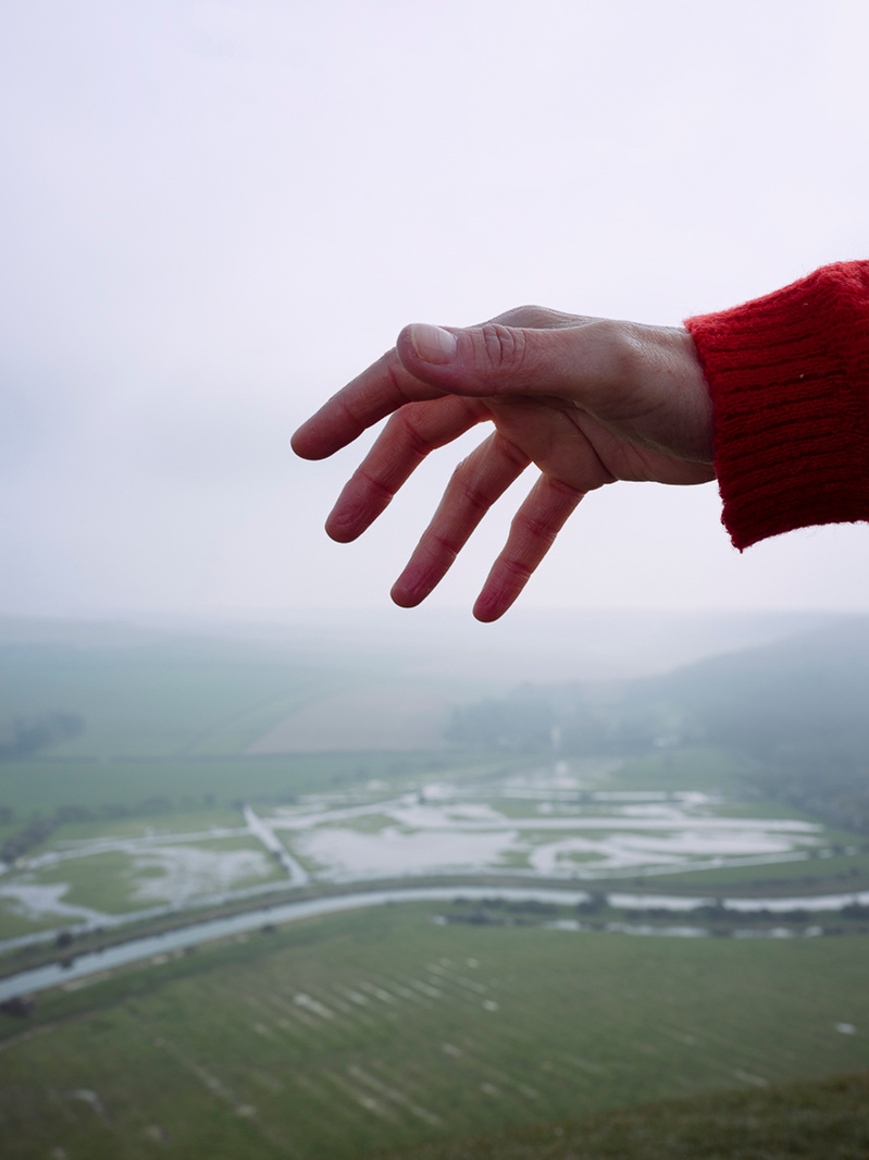 Colour photograph of a hand reaching out above a landscape in the background.