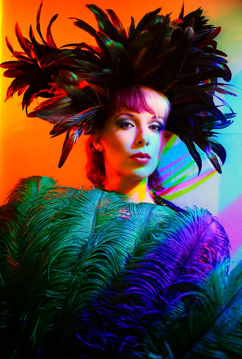 Colour photograph of a young woman wearing a large feather headdress and holding a coloured feather fan in front of her.