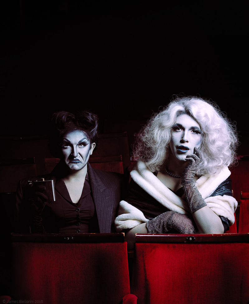 Colour photograph of two drag performers dressed as 1940s Hollywood film stars looking at the camera and sitting in red velvet cinema seats.