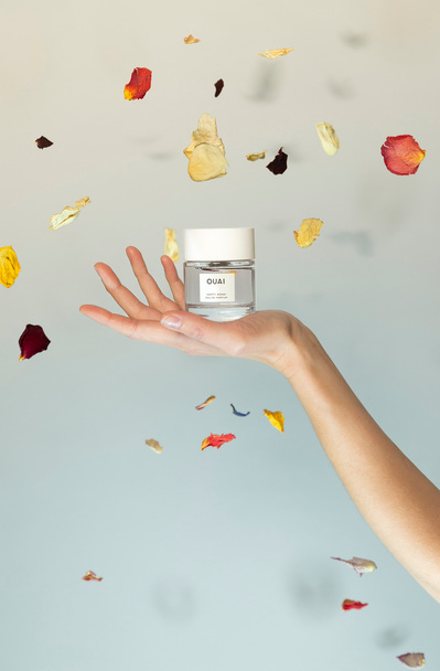Ouai fragrance product shot in model's hand with flower petals falling around it.