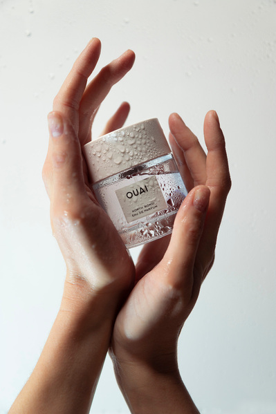 Ouai fragrance bottle held by model's hands against a white backdrop.