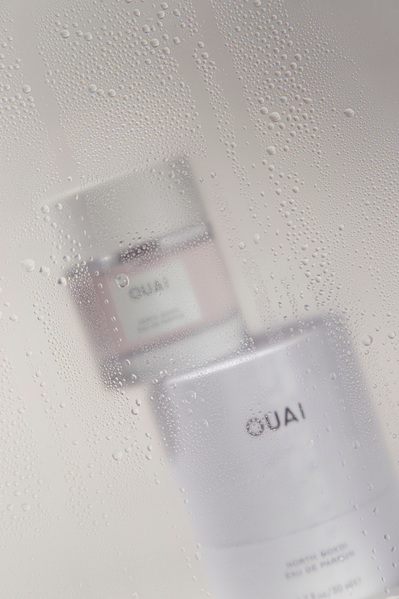 Ouai fragrance bottle sits on top of product packaging behind transparent pane spritzed with water droplets.