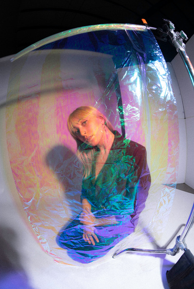 Editorial fashion model in burgundy blazer kneeling behind iridescent cellophane. 