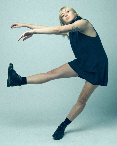Editorial fashion model in a navy tank dress and boots sways to the side balancing on one foot on a white backdrop.