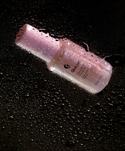 Glossier Future Dew bottle laying on top of a transparent panel covered in water droplets against a black backdrop.