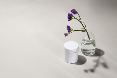 Ouai fragrance packaging sits next to a clear vase with a single purple flower stem on a light grey backdrop.