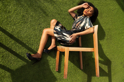 Lifestyle fashion model lays on astroturf in the sun while appearing to sit in a chair that's turned on its side.