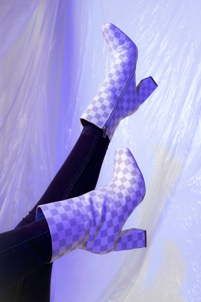 Lifestyle fashion model wearing white and grey checkered ankle boots and black skinny jeans against a purple-tinted plastic backdrop.