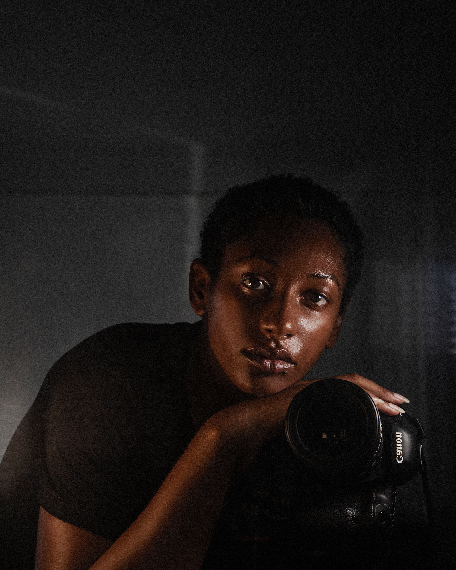 Black woman in black shirt taking a self-portrait with Canon camera in a mirror with dark, ambient, lighting.