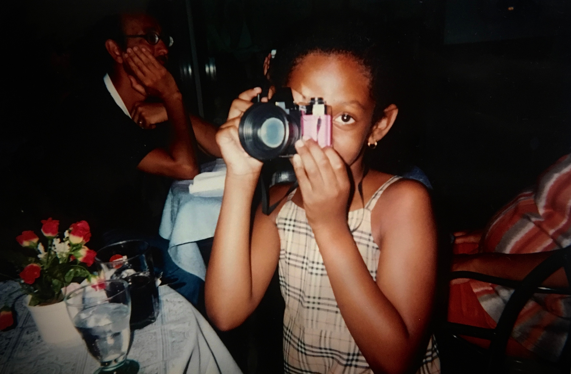 Child in patterned dress holding a pink toy camera up to her face at a restaurant