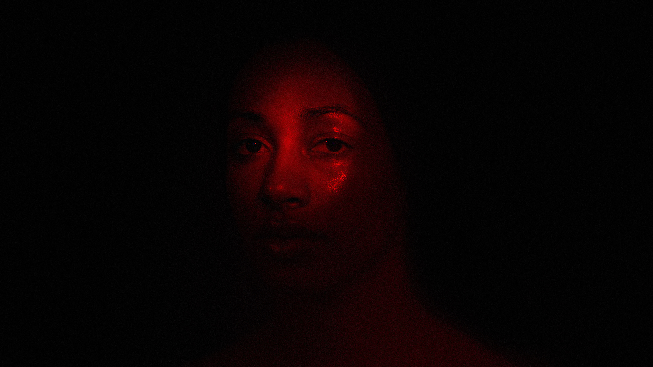 Portrait of a black woman under red light looking at the viewer in front of a black background.
