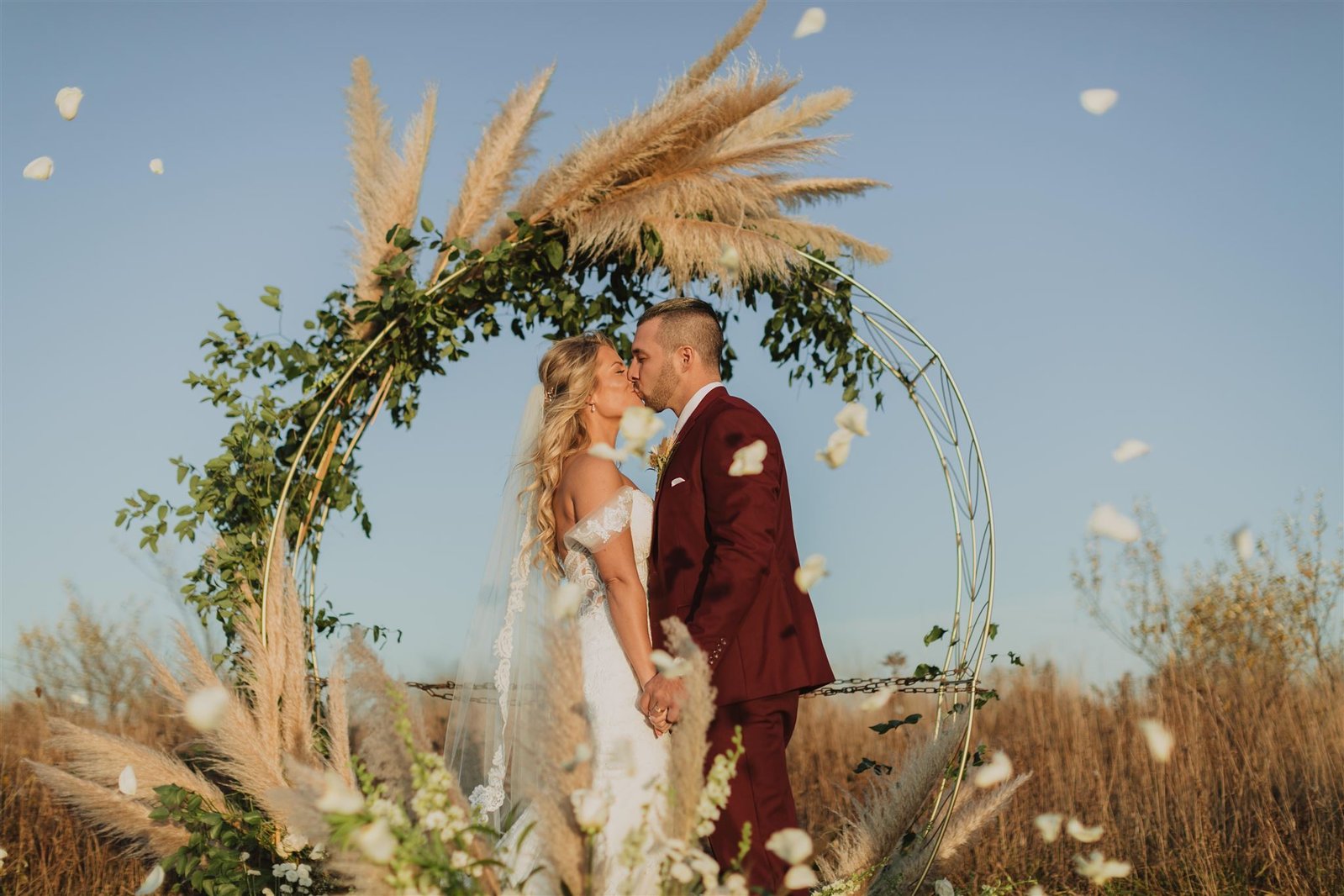bride and groom kissing and holding hands under the arbor with rose petals flying through the air on their wedding day