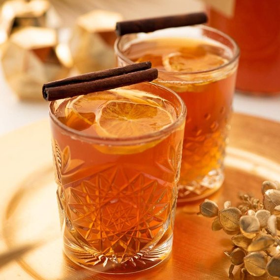 Two glasses of cider sit on a gold tray with a festive props and cinnamon stick garnishes.