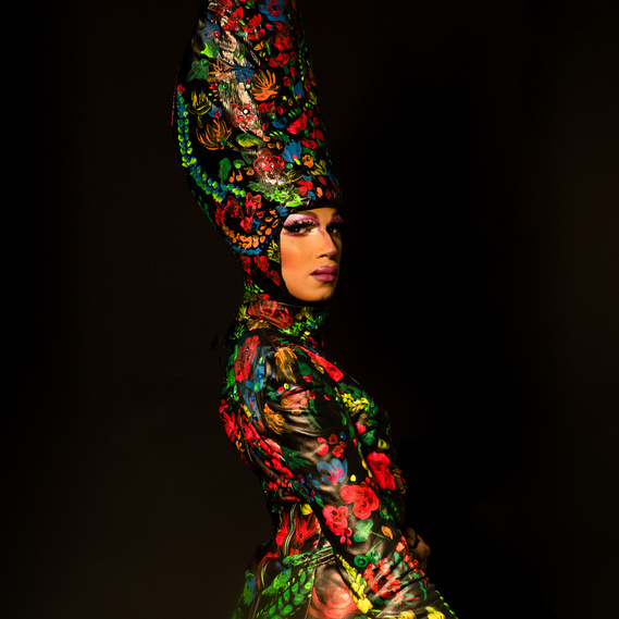 Bombae, a toronto drag queen, stands in front of a black background. She is looking back over her right shoulder. She is wearing a floral painted outfit and elaborate hat,