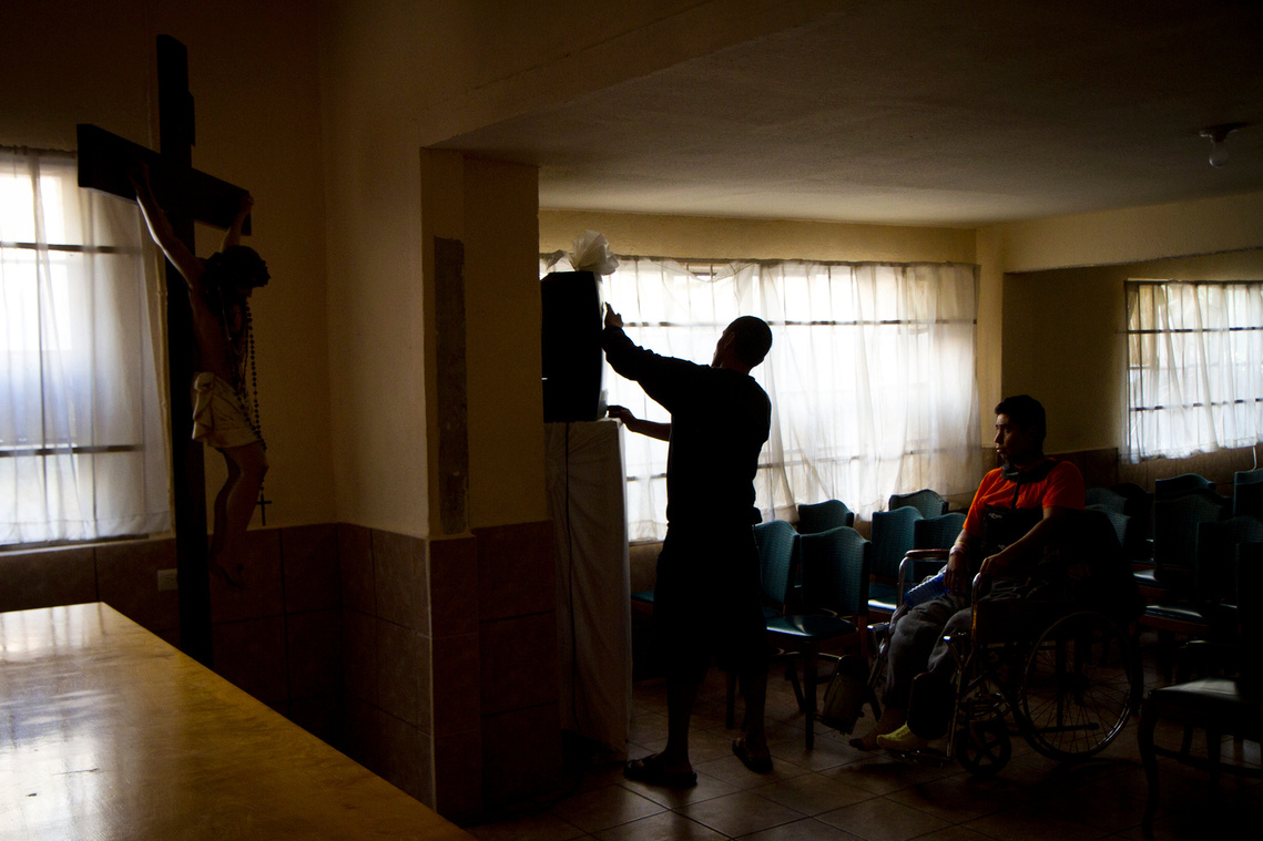 Rodriguez turns on a television in the chapel at Albergue San Juan Bosco for Martinez early in the morning after most migrants at the shelter left for the day. Rodriguez looked after Martinez as much as he could after he arrived at the shelter.