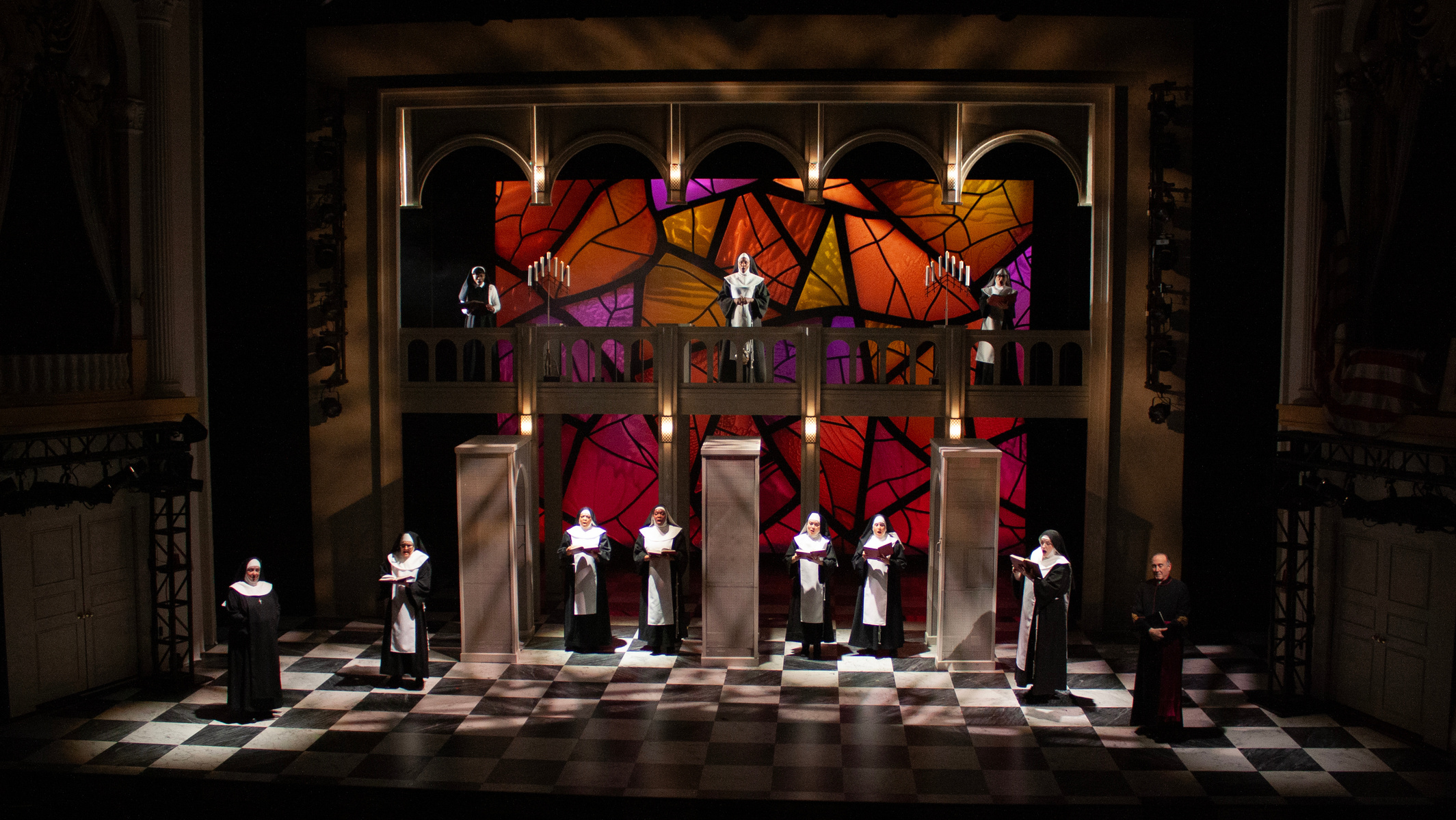 Paige Hathaway's scenic design for "Sister Act", directed by Jeff Calhoun at Ford's Theatre in 2025. The design consists of spinning panels, a large portal with theatrical lights, a checkerboard marble floor, and a backlit stained glass backdrop.