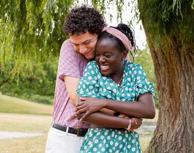 Man hugging woman from behind underneath weeping willow tree. Couple is smiling.