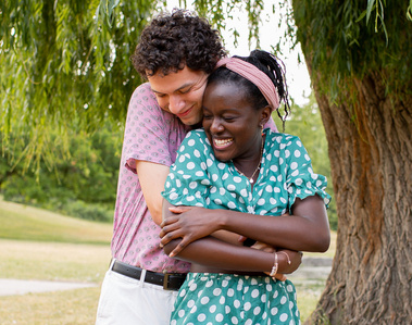 Man hugging woman from behind underneath weeping willow tree. Couple is smiling.