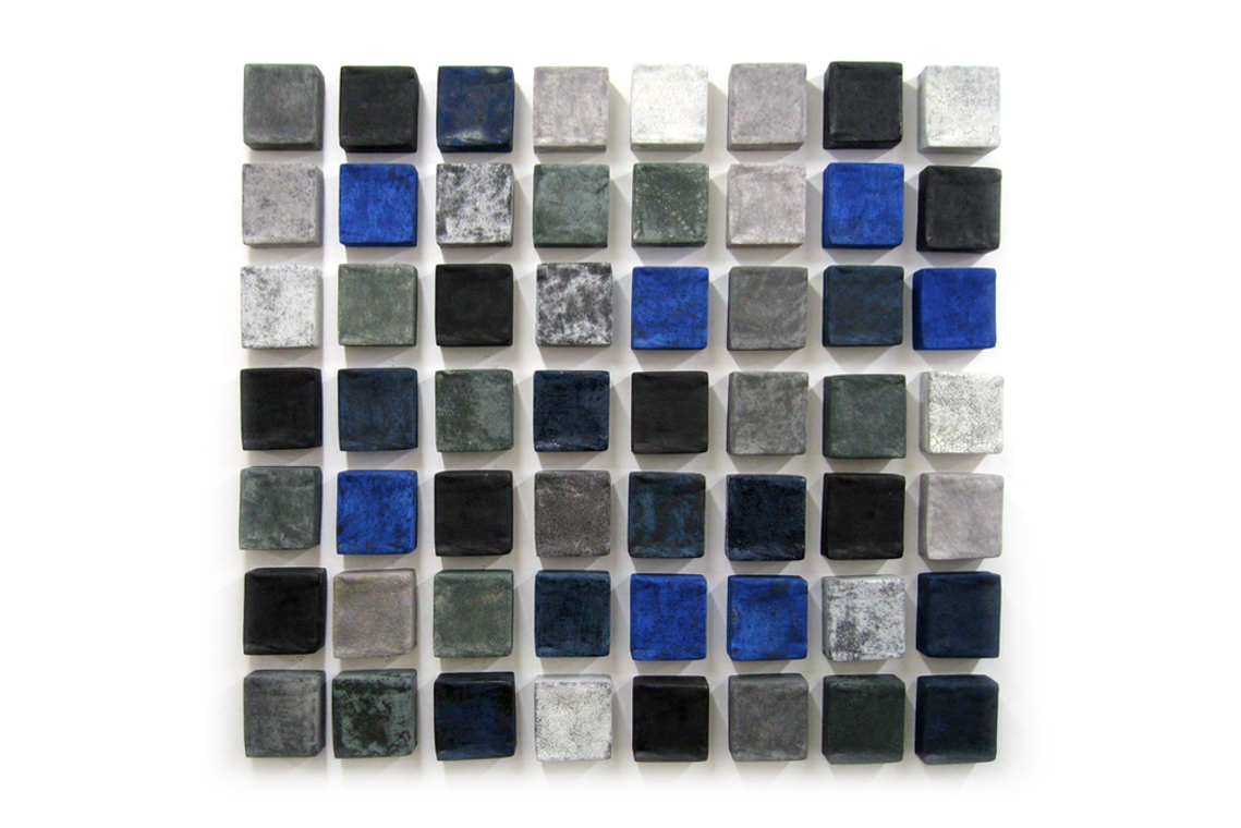 Four-inch ceramic squares that were slip cast by Kathy Erteman make up the Indigo Flush, extended view, architectural tile art. The squares vary in color from Yves Klein blue, grey, and charcoal to white, and are arranged with space between the squares