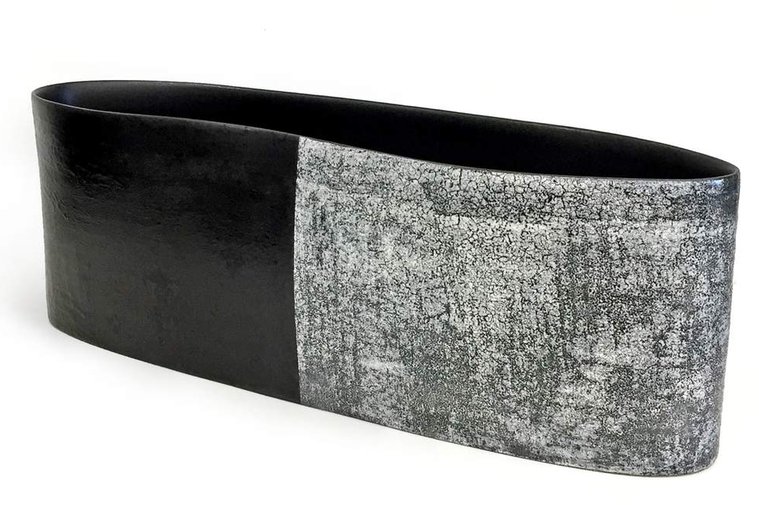 Wheel thrown and altered, Asphalt and Snow 3 by Kathy Erteman is a curved long vessel made of porcelain with a black texture slip and texture white glaze visually dividing the piece of art in half 