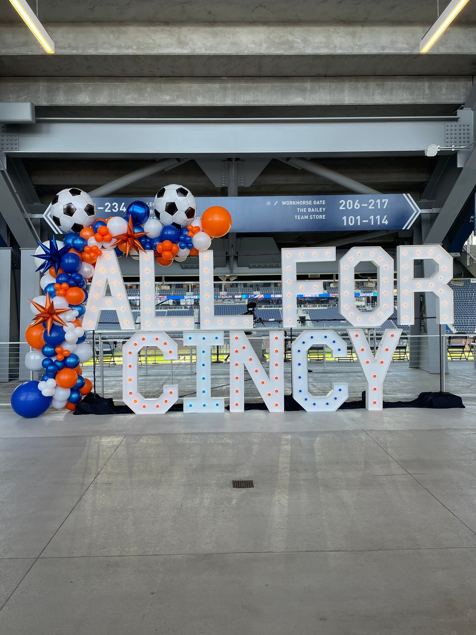 under a stadium are large white light up marquee letters with blue and orange lights and a balloon garland draped over of blue and orange balloons with soccer ball mylar balloons
