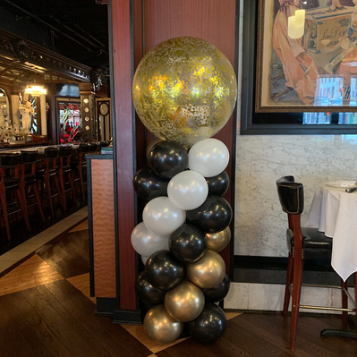 In a restaurant space in front of a wood panel is a spiral balloon column of white black and gold balloons with a large clear balloon with gold confetti inside