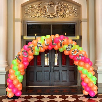 In front of a doorway is a balloon arch of pink green and orange balloons with mylar gold letters over top