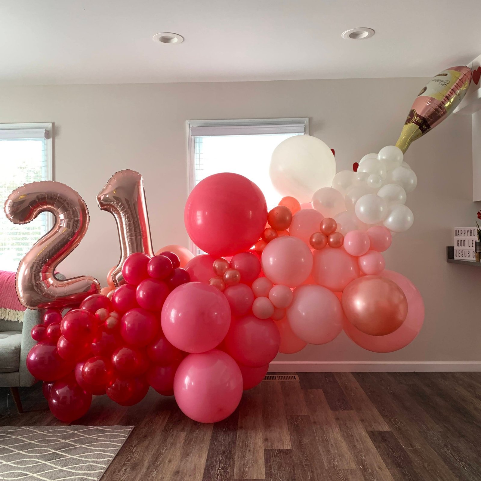In front of a white wall is a large balloon garland of pink and white balloons with 21 number mylar balloons and a champagne bottle balloon