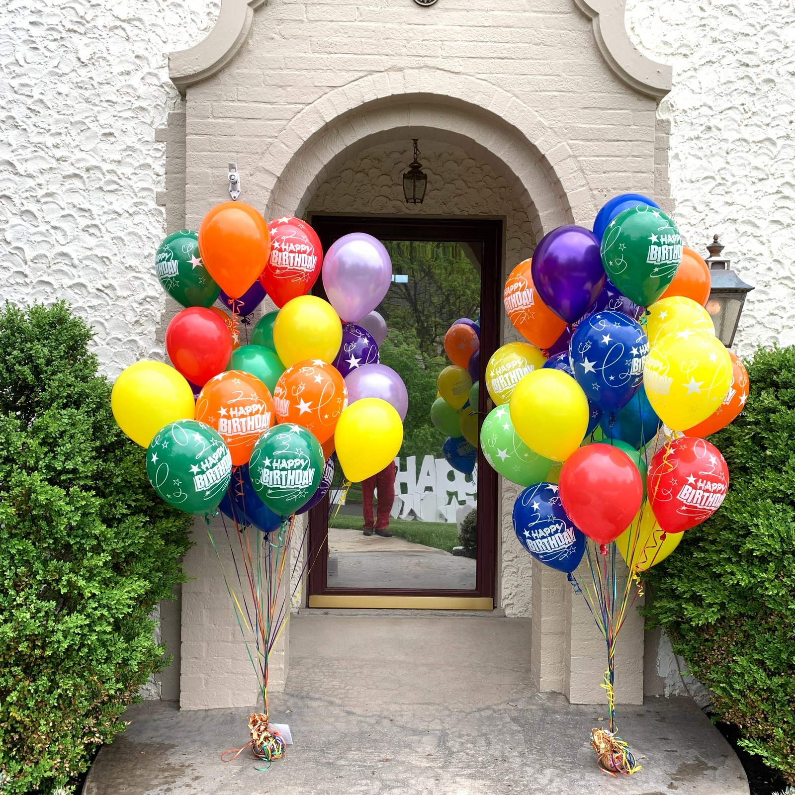 In front of an entryway to a white brick home are two large helium balloon bouquets of red yellow orange blue green and purple with happy birthday text on them