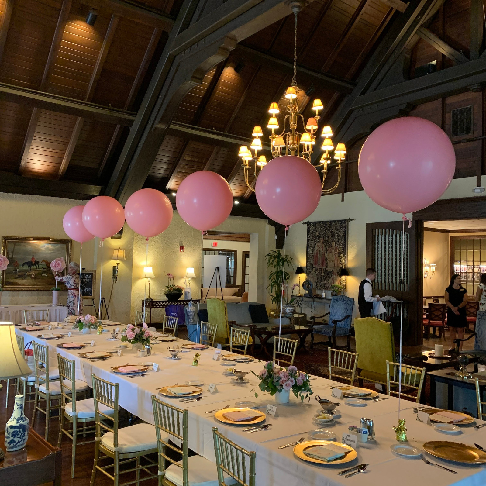 In an event space is a long table with a white tablecloth and place settings with centerpieces of pink helium balloons